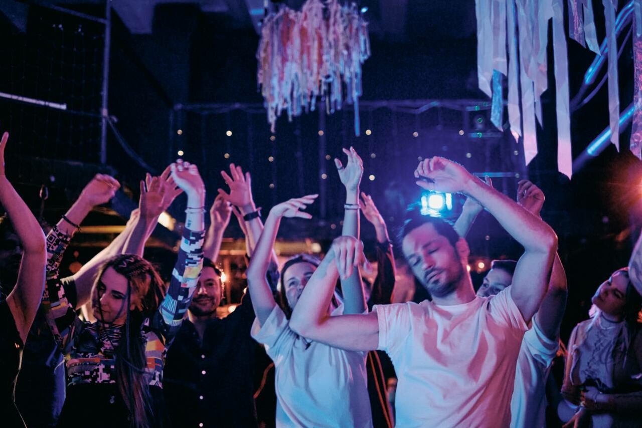 Арендовать Лес. Театральный лофт с верандой, сценой и вторым ярусом. Внутреннее пространство с музыкой и танцующими людьми, разноцветное освещение, включающее синие и фиолетовые тона, подвесные декоративные элементы на потолке, плотная атмосфера.