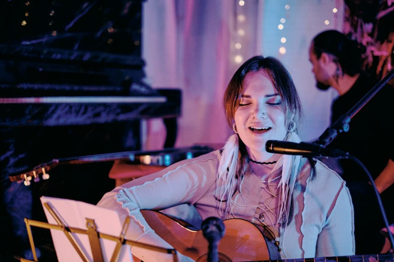 Арендовать Лес. Театральный лофт с верандой, сценой и вторым ярусом. Музыкальное пространство с деревянным пианино и стулом, натяжной потолок украшен светодиодными гирляндами. Человек играет на акустической гитаре перед микрофоном, перед ним нотная стойка. Освещение мягкое, тёплых тонов.