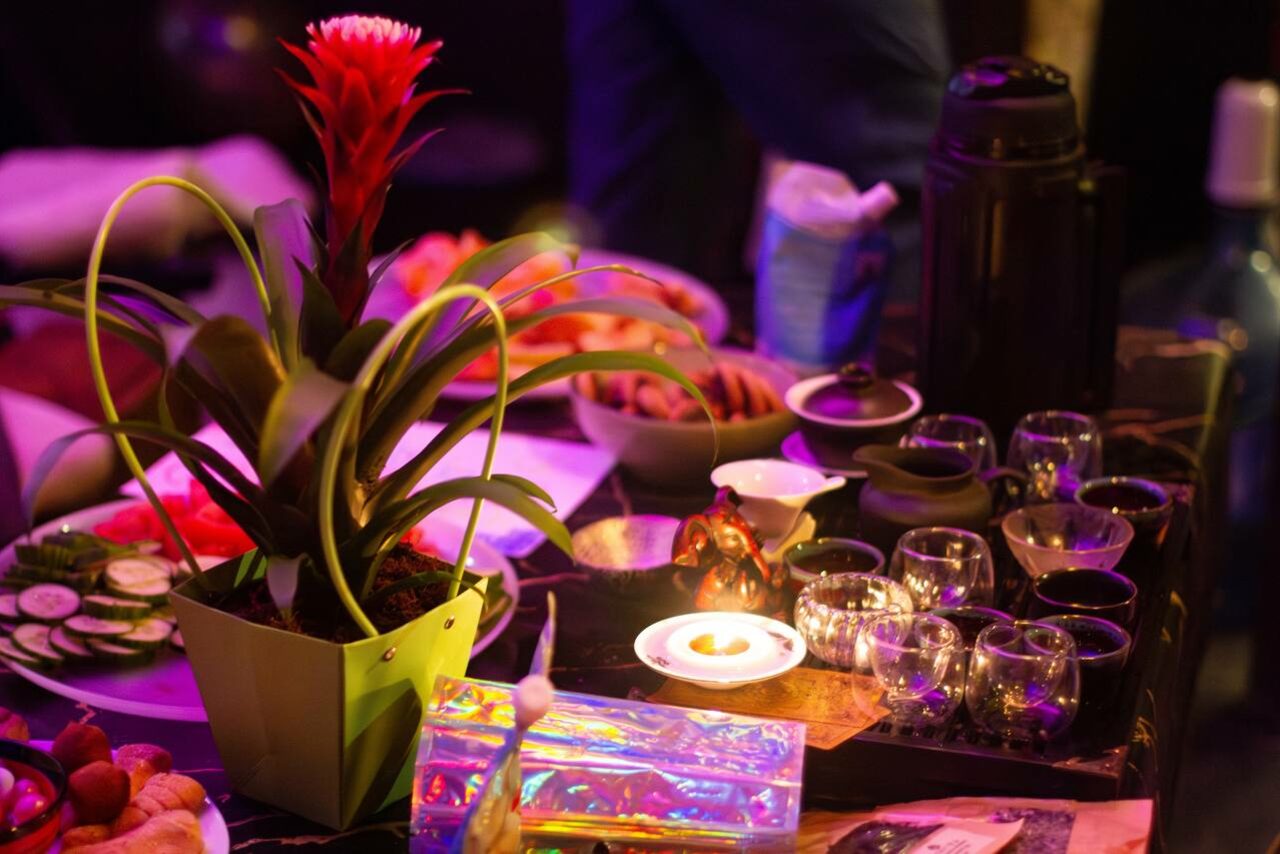 Арендовать Лес. Театральный лофт с верандой, сценой и вторым ярусом. Стол с декоративным растением в зеленом горшке, напитками в стеклянной и керамической посуде, разноцветными упаковками, свечой на подставке. Освещение теплое, розово-оранжевое. На фоне видны различные упаковки и контейнеры.