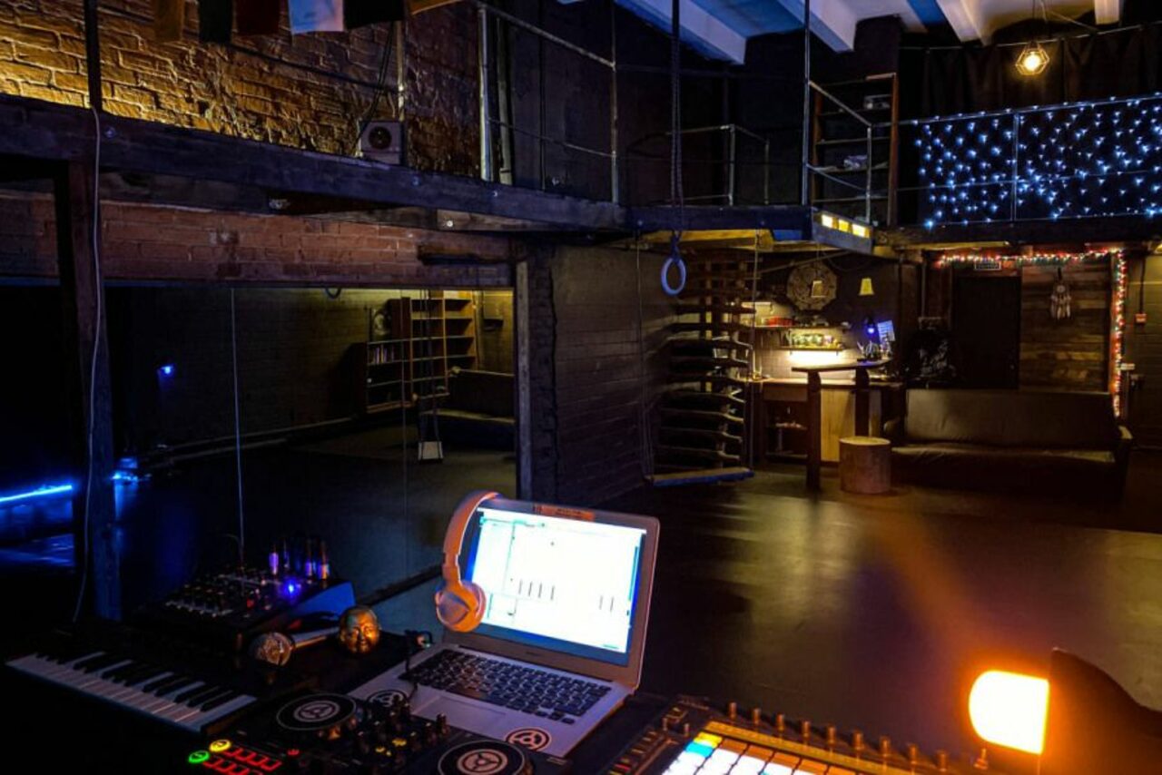 Арендовать Лес. Театральный лофт с верандой, сценой и вторым ярусом. Лофт-пространство с кирпичными стенами и антресолью. На переднем плане диджейский пульт с ноутбуком и наушниками. Подвесные светильники, звуковое оборудование. Освещение приглушенное, синие и теплые оттенки. Столы, стойки и платформа. Светодиодные гирлянды.