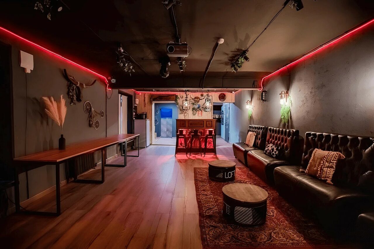 Арендовать Пространство для любых мероприятий с верандой и грилем. Лофт-пространство с деревянным полом, кожаным диваном, деревянными столами. Красная подсветка на стенах, потолок с прожекторами. На стенах декор, подушки на диване. Барная стойка с напитками, два круглых стола. Тёмные стены.