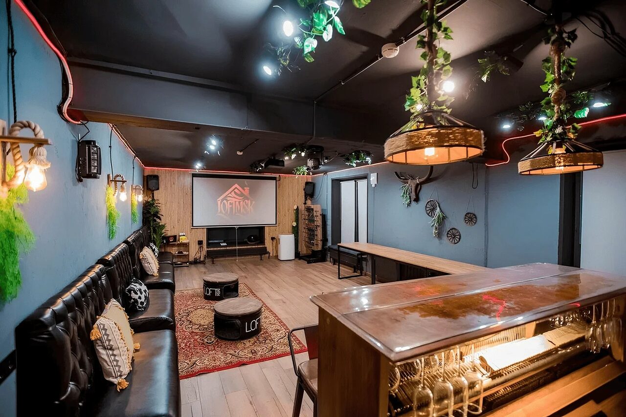 Арендовать Пространство для любых мероприятий с верандой и грилем. Лофт-пространство с барной стойкой, длинным столом, диванами, пуфами, проектором, экранами. Потолочные светильники, декоративные элементы и растения. Деревянный пол, стены синие, потолок темный.