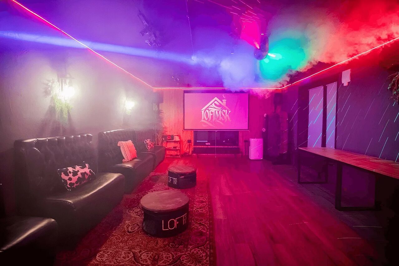 Арендовать Пространство для любых мероприятий с верандой и грилем. Лофт-пространство с черными кожаными диванами, маленькими столиками, большим экраном и надписью "LOFT". Темное освещение с цветными световыми эффектами, дым. Деревянный пол и стол, ковёр. Светильники на стенах.