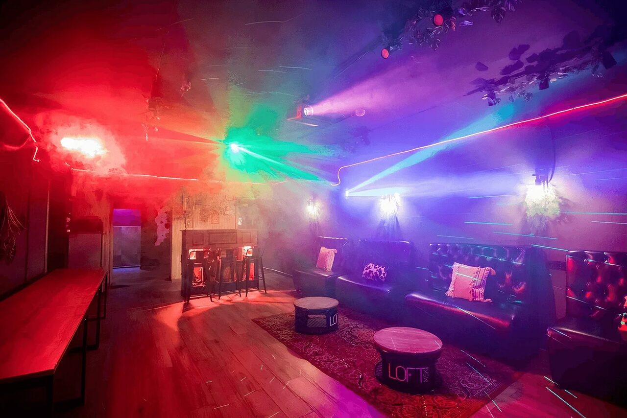Арендовать Пространство для любых мероприятий с верандой и грилем. Лофт-пространство с длинным столом и диваном у стены. На полу ковры, в центре круглые столики. Освещение разноцветными прожекторами, стены украшены растениями. Потолок с лазерами. Красные и синие оттенки, надпись "LOFT" на столах.