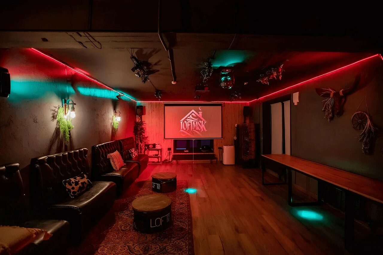 Арендовать Пространство для любых мероприятий с верандой и грилем. Помещение для мероприятий с кожаными диванами вдоль стены, деревянным полом и большим экраном. Потолок украшен неоновыми светильниками, создающими атмосферное красное и зеленое освещение. Присутствуют декорации в виде растений на стенах.