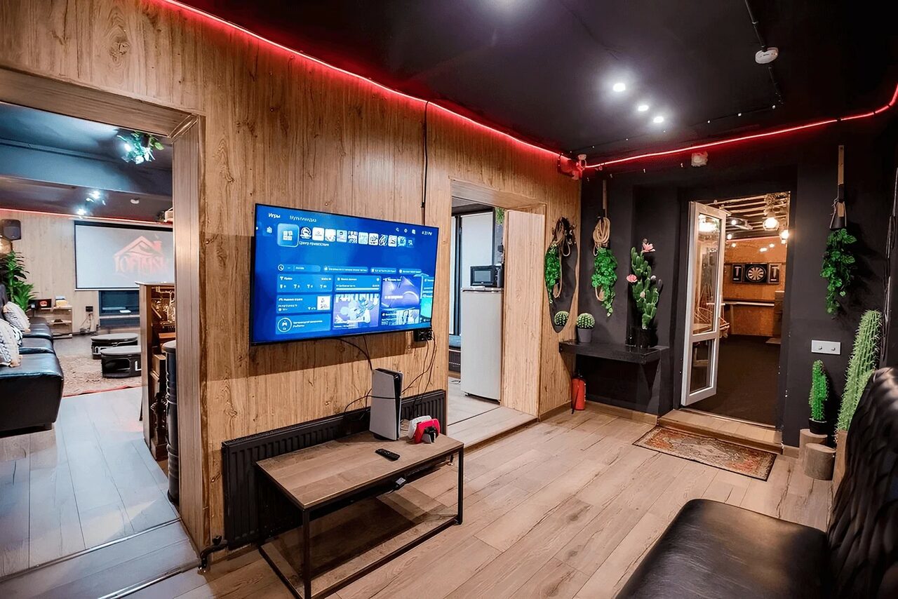 Арендовать Пространство для любых мероприятий с верандой и грилем. Меблированное пространство с деревянными панелями и черным потолком, декоративная светодиодная подсветка. На стене закреплен телевизор над черным радиатором. В комнате широкоэкранная дверь, проходящая в соседнюю зону. На полках декоративные растения.