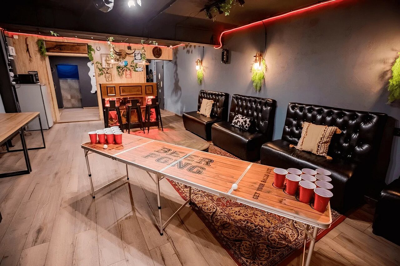 Арендовать Пространство для любых мероприятий с верандой и грилем. Пространство с меблировкой для отдыха: длинный стол для игр с красными пластиковыми стаканами, черные кожаные диваны с декоративными подушками, деревянный пол, растения на стенах, деревянный шкаф, настенные светильники, неоновая подсветка.