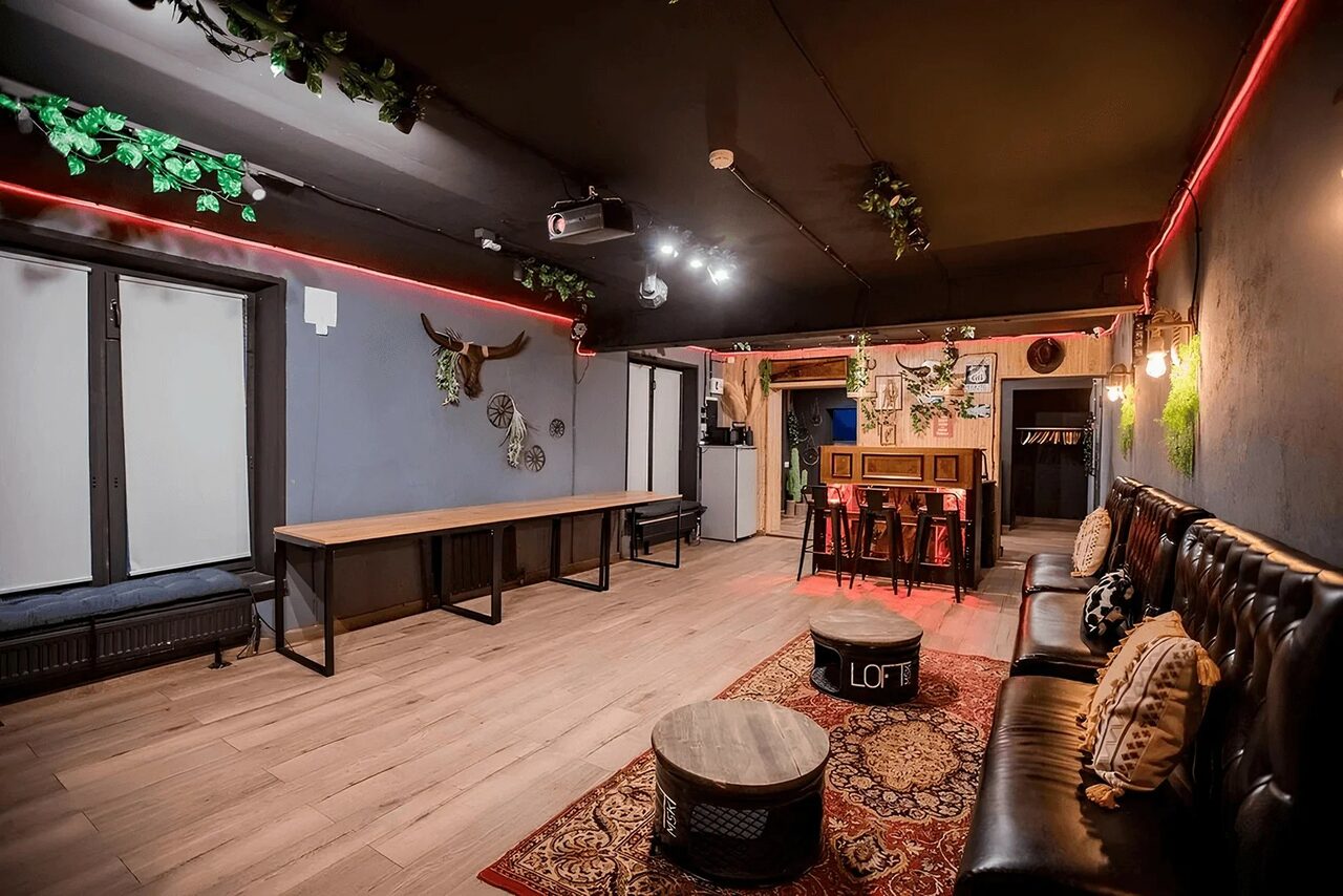 Арендовать Пространство для любых мероприятий с верандой и грилем. Пространство с деревянным полом, стены темные, черный потолок с растениями, длинный стол у окна, диваны с подушками, кофейные столики, ковёр, освещение комбинированное, неоновая подсветка, проекционное оборудование, картины на стенах.