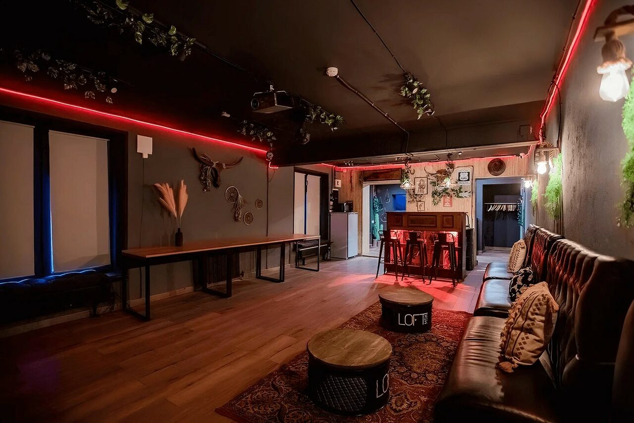 Арендовать Пространство для любых мероприятий с верандой и грилем. Лофт-пространство с темными стенами и потолком. В центре комнатные диваны и столики на ковре, у стены длинный стол. Настенные декорации и подсветка красного цвета. Потолочные подвесы с зелеными элементами. Доска для написей, проектор.