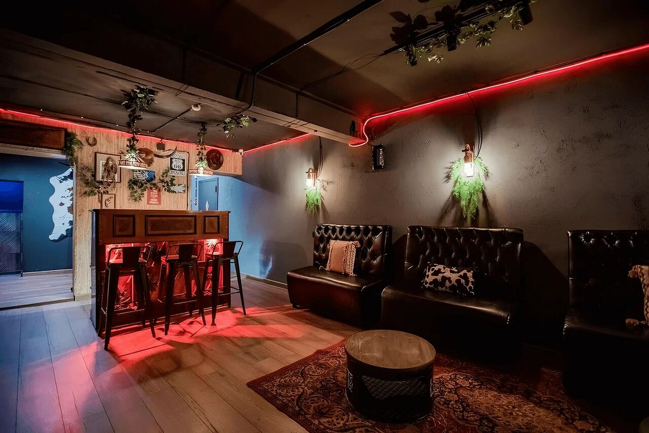 Арендовать Пространство для любых мероприятий с верандой и грилем. Тёмное помещение в стиле лофт, с неоновым красным освещением. Темные кожаные диваны, деревянный бар с высокими табуретами, растения на стенах, ковёр на полу. Стены темно-серые и деревянные, потолок с декором из зелени.