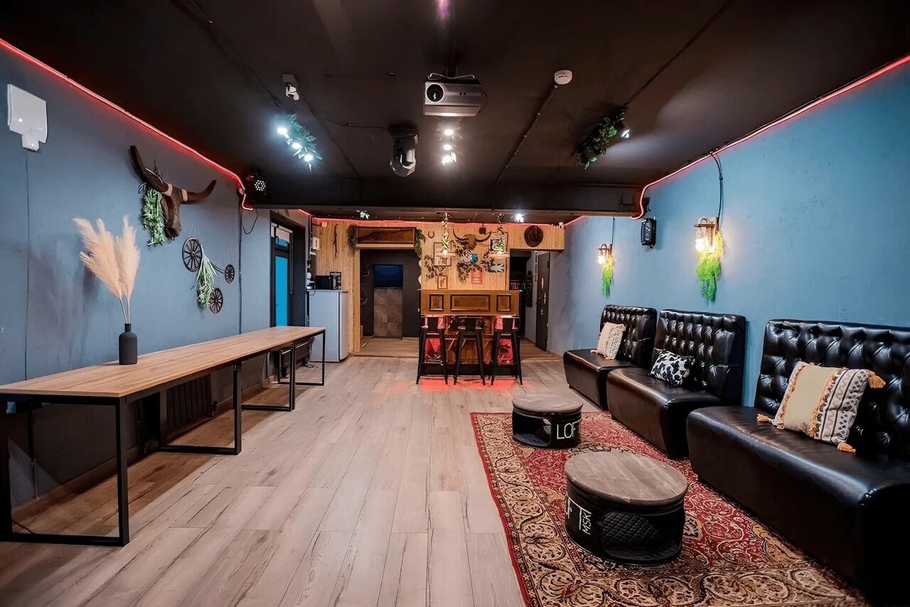 Арендовать Пространство для любых мероприятий с верандой и грилем. Пространство с синими стенами и черным потолком, оборудованное проектором и трековыми светильниками. Слева длинный деревянный стол. Справа кожаные диваны и барные стулья, журнальные столики. Красный ковёр на полу. Настенные декорации и зелень.