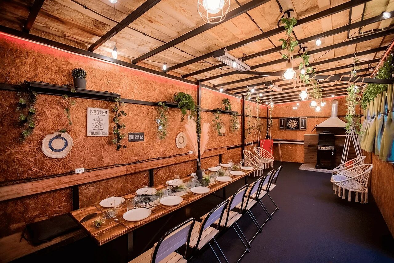 Арендовать Пространство для любых мероприятий с верандой и грилем. Пространство в деревянной обшивке, длинный стол с посудой и стульями, подвесные лампы, украшения из зелени, дартс на стене, зона с мангалом, потолок из дерева и металла, боковые стены с декором, кресла-подвесы, разноцветные шторы.