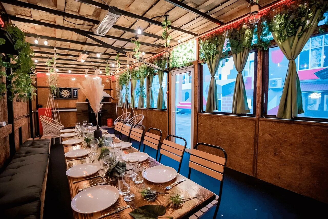 Арендовать Пространство для любых мероприятий с верандой и грилем. Помещение с деревянными стенами и потолком, продолговатый стол с сервировкой для банкета на много гостей. Металлические и деревянные стулья, зелёные шторы и декор из растений. Настенные светильники, дартс на стене, подвесные кресла и диван вдоль стены.