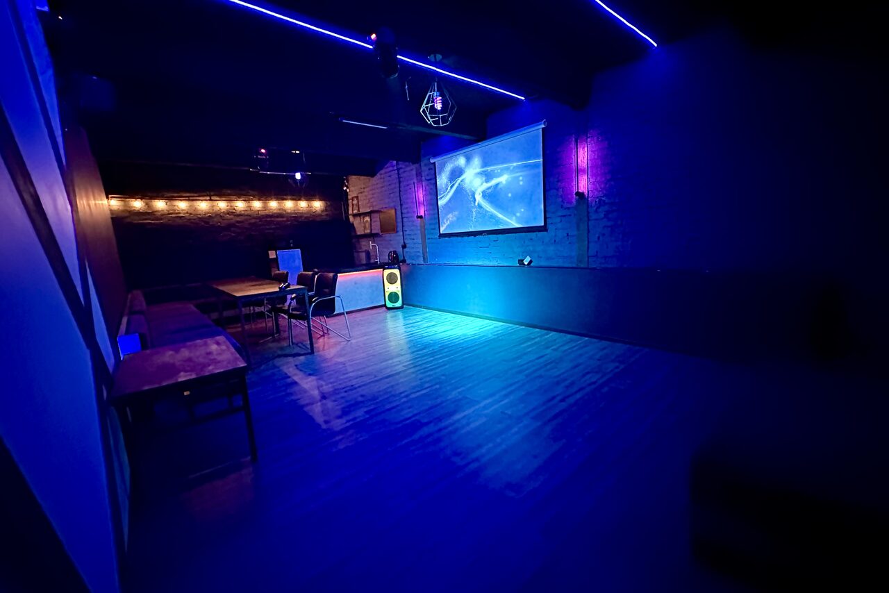 Арендовать Kahovka loft (Каховка Лофт). Неоновый лофт-зал. Комната с темными стенами, деревянным полом, столами и стульями слева. Напротив висит экран с проектором. На стенах неоновое освещение и светящиеся декоративные элементы. В углу стоит колонка.