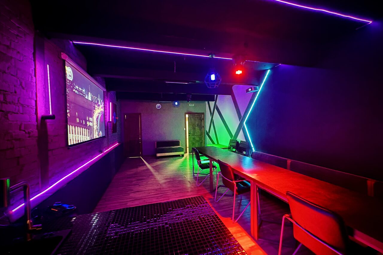 Арендовать Kahovka loft (Каховка Лофт). Неоновый лофт-зал. Темное помещение с неоновыми акцентами. Справа длинный деревянный стол с разноцветными стульями. На стенах и потолке неоновые ленты фиолетового и зеленого цветов. Экран на левой стене, деревянный пол и двери.