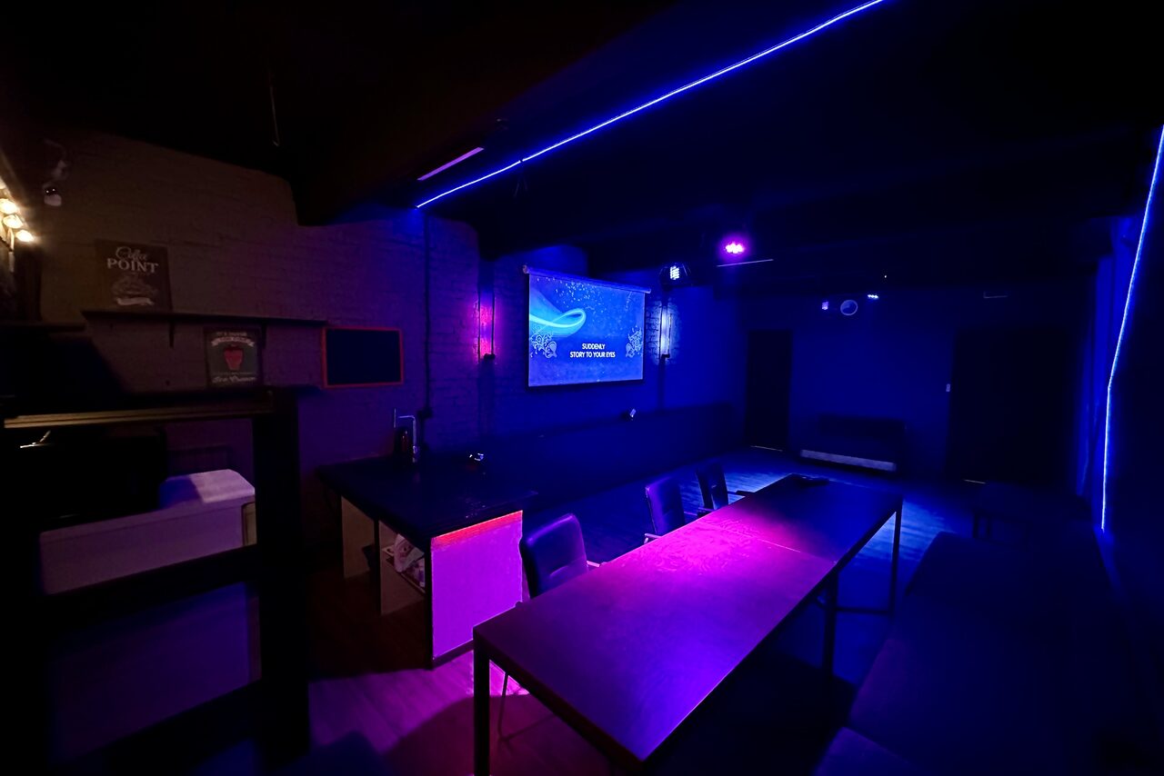 Арендовать Kahovka loft (Каховка Лофт). Неоновый лофт-зал. Тёмное помещение с длинным столом и стульями. Наличие фиолетовой и синей подсветки. На стене экран с изображением. Стеллаж с коробками справа, кирпичная стена слева. Современное оборудование и мебель.