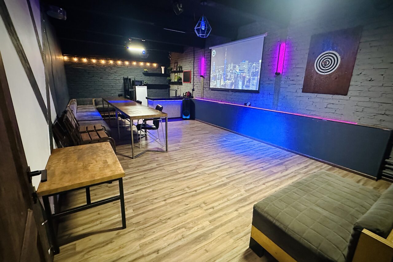 Арендовать Kahovka loft (Каховка Лофт). Неоновый лофт-зал. Пространство с деревянным полом, кирпичными стенами и мебелью из светлого дерева; длинный стол с рядом стульев; диваны вдоль стен; на стене проектор с изображением городской панорамы; разноцветная подсветка по периметру.