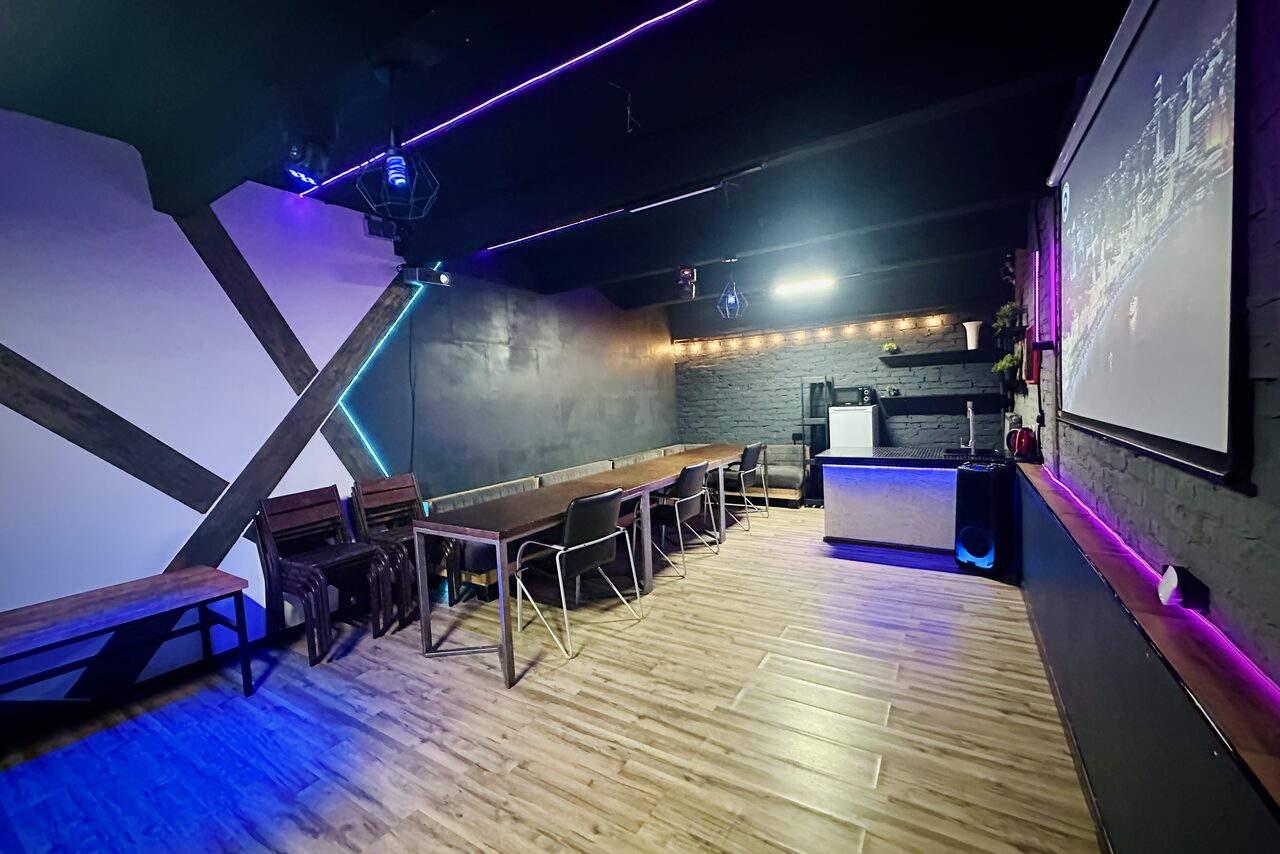 Арендовать Kahovka loft (Каховка Лофт). Неоновый лофт-зал. Пространство с ламинированным полом, тёмными стенами и живописным контрастным потолком с неоновыми светильниками. Несколько столов и стульев, проекторная стена и барная стойка на заднем плане. Освещение теплое, атмосфера современная.