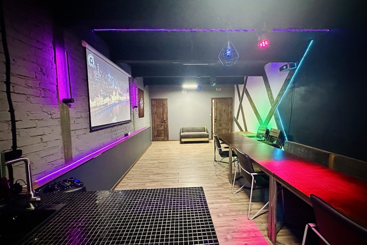 Арендовать Kahovka loft (Каховка Лофт). Неоновый лофт-зал. Пространство в индустриальном стиле с кирпичными стенами, барной стойкой, стульями и диваном. Светодиодное освещение разноцветное, ламинат на полу. На стене проектор с изображением ночного города. Две деревянные двери.