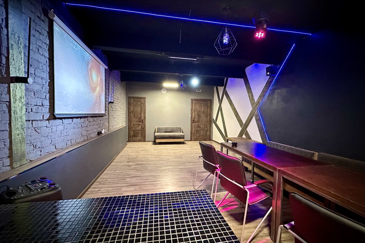 Арендовать Kahovka loft (Каховка Лофт). Неоновый лофт-зал. Помещение с кирпичными стенами, проектором, столом и стульями, мягким диваном. Потолок черный с подвесными лампами и светодиодной подсветкой. Паркетный пол, двери деревянные. Стены серые с декоративными элементами. Настенное изображение.