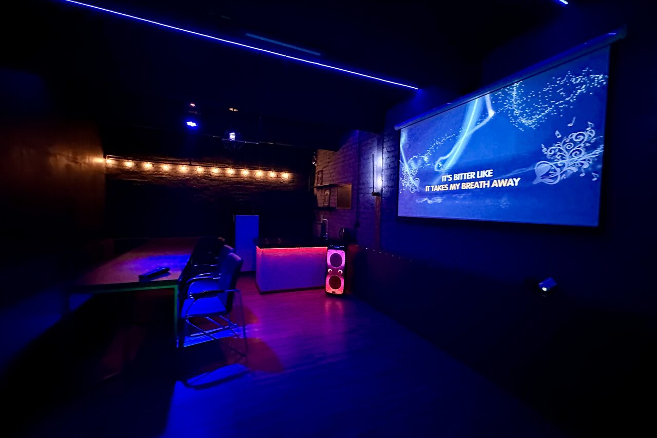 Арендовать Kahovka loft (Каховка Лофт). Неоновый лофт-зал. Тёмное помещение с проектором на стене, столом и стульями. Освещение с помощью светодиодных лент и светильников. Стены чёрные, поверхность с кирпичной текстурой. Напольное покрытие светло-коричневое, экран отображает анимацию и текст.