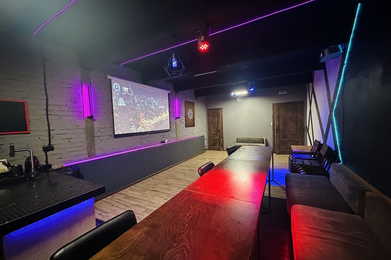 Арендовать Kahovka loft (Каховка Лофт). Неоновый лофт-зал. Просторное помещение с длинным деревянным столом и мягкими диванами по бокам. На стене проектор, кирпичная кладка, деревянные двери. Яркое цветное освещение вдоль потолка и пола. Открытая кухня барного типа слева.