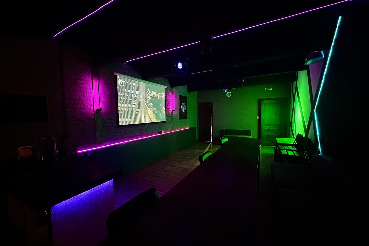 Арендовать Kahovka loft (Каховка Лофт). Неоновый лофт-зал. Темное помещение с неоновым освещением, фиолетовыми и зелеными лампами. Столы и стулья вдоль стен, проектор на стене, показывающий изображение. Пол деревянный. Пространство подходит для встреч или мероприятий.