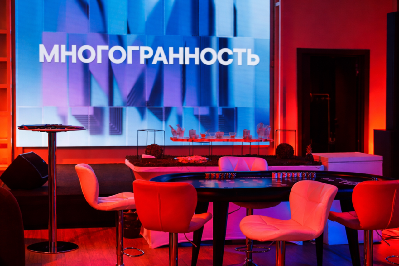 Арендовать Дворец Культур. Пространство с круглыми стульями, картежным столом, высокой барной стойкой. Задник с крупным текстом "МНОГГРАННОСТЬ" на экране сине-фиолетового оттенка. Освещение тёплое и красное. Деревянный пол.