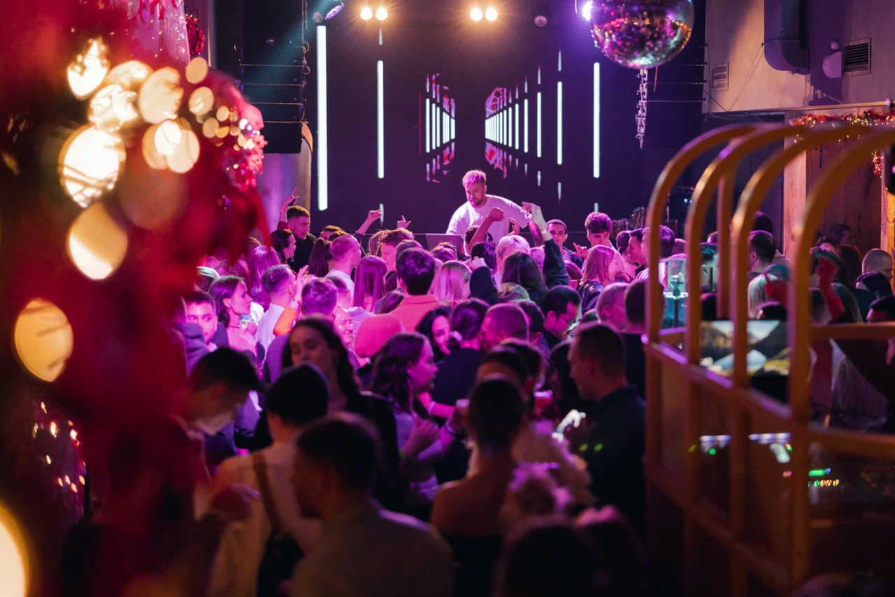 Арендовать Бар StereoPeople (СтереоПипл). Ночной клуб с большим количеством людей на танцполе. На заднем фоне диджей за пультом. Цветное освещение: розовые и фиолетовые оттенки. Декор из гирлянд и подвесных шаров. На стене светодиодная вертикальная инсталляция.