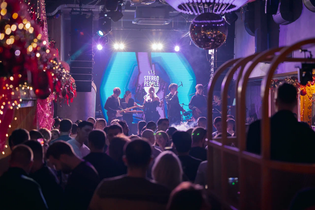Арендовать Бар StereoPeople (СтереоПипл). Концертное пространство с живой музыкой, большой экран на сцене, группа музыкантов, темное освещение, подвесной светильник в виде дискошара, плотная толпа людей, праздничные декорации с гирляндами и шарами.