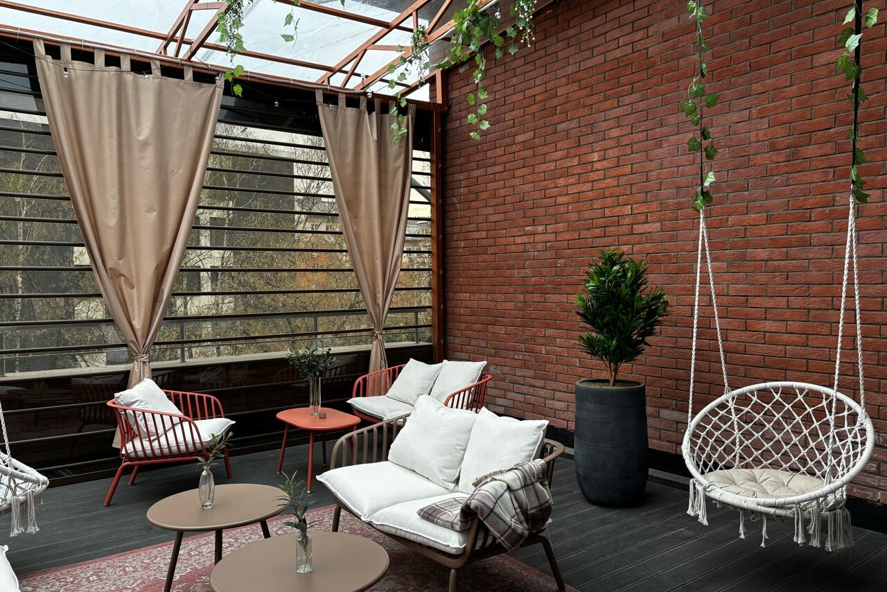 Арендовать Лофт Loftsinoby (Лофтсиноби)  для мероприятий. Терраса с красными кирпичными стенами и стеклянной крышей. Мебель: белые диваны, красные стулья, плетёное кресло-кокон. На полу тёмно-коричневые доски, есть ковры. Декор: зелёные растения и подвесные гирлянды. Шторы бежевого цвета.
