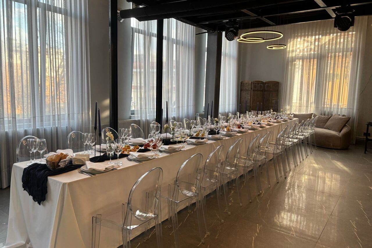 Арендовать Лофт Loftsinoby (Лофтсиноби)  для мероприятий. Просторное помещение с длинным столом, накрытым белой скатертью. Прозрачные стулья, сервировка из бокалов и тарелок. Настенные светильники, большие окна с белыми занавесками, светлые стены, оттенки серого и коричневого.