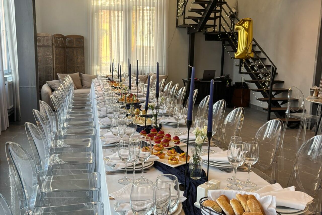 Арендовать Лофт Loftsinoby (Лофтсиноби)  для мероприятий. пространство для мероприятий; длинный обеденный стол с белой скатертью, прозрачные стулья, сервировка бокалами и столовыми приборами; свечи в подсвечниках; тарелки с угощениями; металлическая лестница; воздушный шар в форме цифры; яркие окна с занавесками.