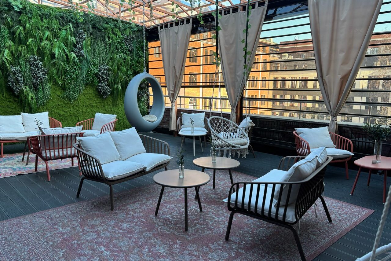 Арендовать Лофт Loftsinoby (Лофтсиноби)  для мероприятий. Открытая терраса с панорамными окнами, плетеными креслами и диванами с подушками. На полу ковёр с растительным орнаментом. Стены украшены зелеными растениями. Столики из светлого дерева. Светлая штора. Вид на здание через окно.