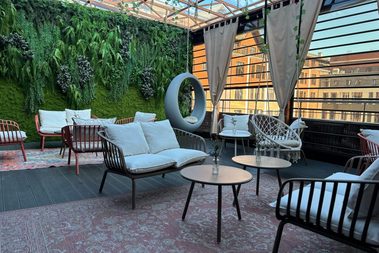 Арендовать Лофт Loftsinoby (Лофтсиноби)  для мероприятий. Открытая терраса с деревянным полом и вертикальной стеной из зелени. Установлены диваны и кресла с подушками, круглый подвесной стул, круглые столы. Пространство украшено занавесками и ковром. Полупрозрачная крыша и обилие естественного света.