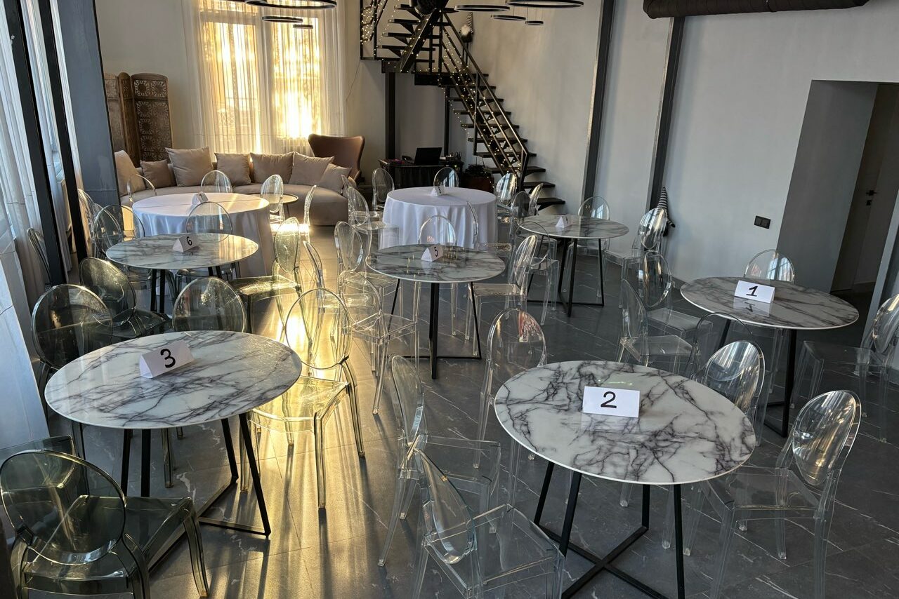 Арендовать Лофт Loftsinoby (Лофтсиноби)  для мероприятий. Просторное помещение с высокими потолками и лестницей. Мраморные круглые столики с прозрачными стульями, на каждом столе номер. Мягкий диван у стены с большими окнами и светлыми шторами. Полы серого цвета. Освещение дневное.