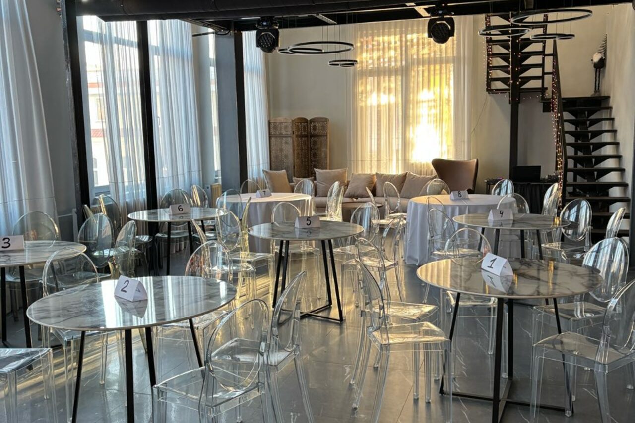 Арендовать Лофт Loftsinoby (Лофтсиноби)  для мероприятий. Современное пространство с несколькими круглыми столами и прозрачными стульями. На столах пронумерованные карточки. Большие окна с полупрозрачными шторами, диван с подушками в дальнем углу. Винтовая лестница, светлое освещение, темный пол.