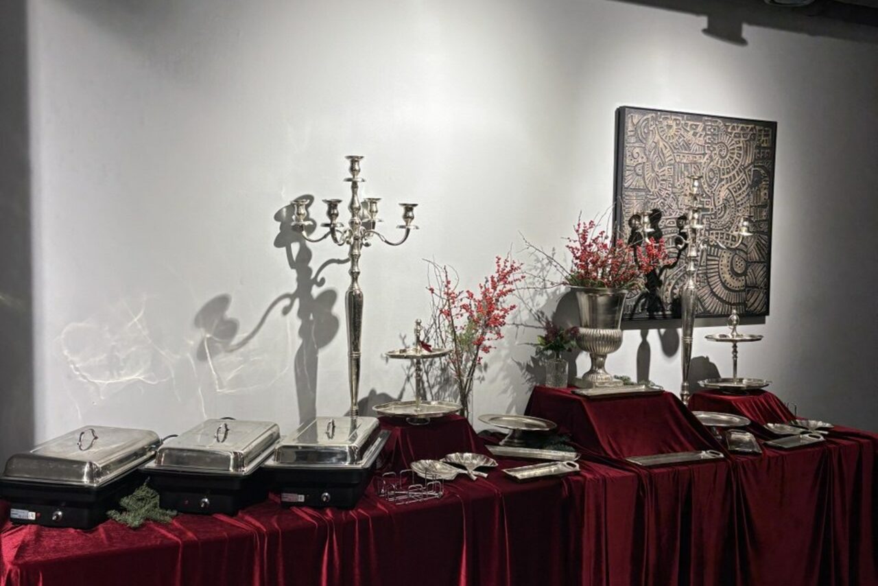 Арендовать Лофт Loftsinoby (Лофтсиноби)  для мероприятий. Стол с бархатной красной скатертью, на столе металлические подогреваемые контейнеры для еды, канделябр, декоративные вазы с ветками и ягодами, сложенные тарелки, фоновая стена с картиной, спокойное освещение.
