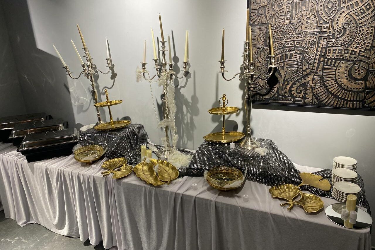 Арендовать Лофт Loftsinoby (Лофтсиноби)  для мероприятий. Стол для фуршета с белой скатертью, украшенный серебристыми подсвечниками с зажженными свечами. На столе золотые блюда, тарелки, стаканы и серебристая ткань. Слева металлические контейнеры для еды. На стене темная картина с узором.