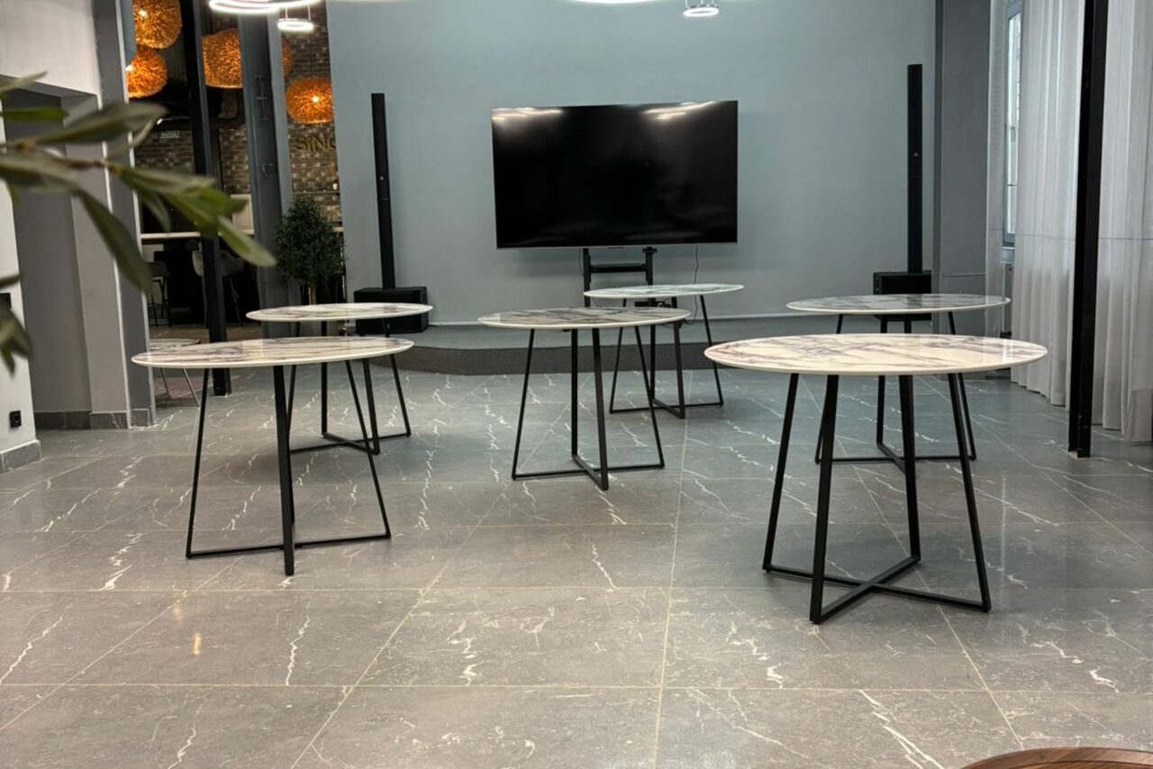 Арендовать Лофт Loftsinoby (Лофтсиноби)  для мероприятий. Современное пространство с серым полом и стенами, несколькими круглыми столами на металлических ножках, большой телевизор на стене, светильники с оранжевым светом, прозрачные занавески на окнах, элементы растений на заднем плане.