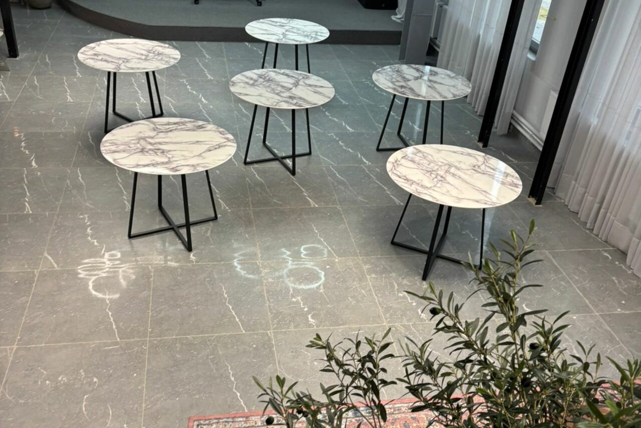 Арендовать Лофт Loftsinoby (Лофтсиноби)  для мероприятий. Пространство с шестью круглыми столами на металлических ножках с мраморными столешницами. Пол из светло-серой плитки с узором. Большие окна с полупрозрачными белыми занавесками. Растение в правом нижнем углу.