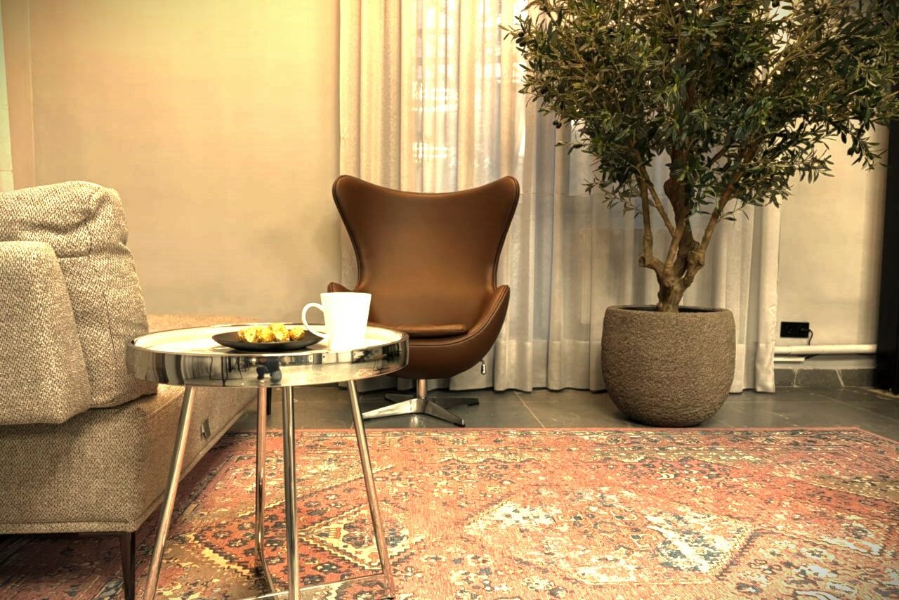 Арендовать Лофт Loftsinoby (Лофтсиноби)  для мероприятий. Гостиная с креслом и диваном, круглый столик с кружкой и тарелкой, большое комнатное растение в горшке, прозрачные шторы, ковер на полу с узором, стены светлого цвета.