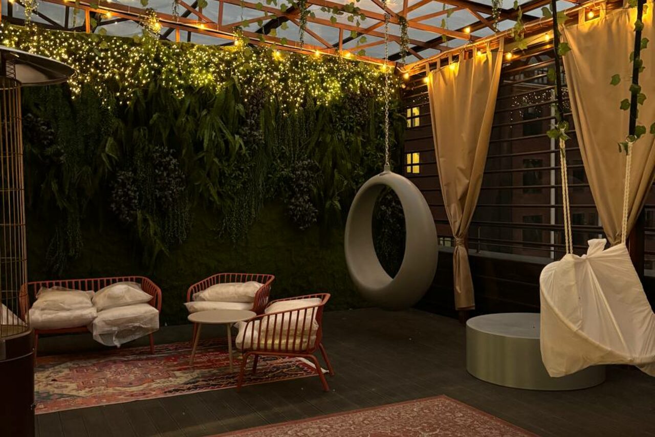 Арендовать Лофт Loftsinoby (Лофтсиноби)  для мероприятий. Открытая терраса с деревянным настилом, мебель из ротанга с подушками, подвесное кресло, мягкое освещение от гирлянд, вертикальное озеленение на стенах, защитные шторы, стол в форме цилиндра, два ковра.