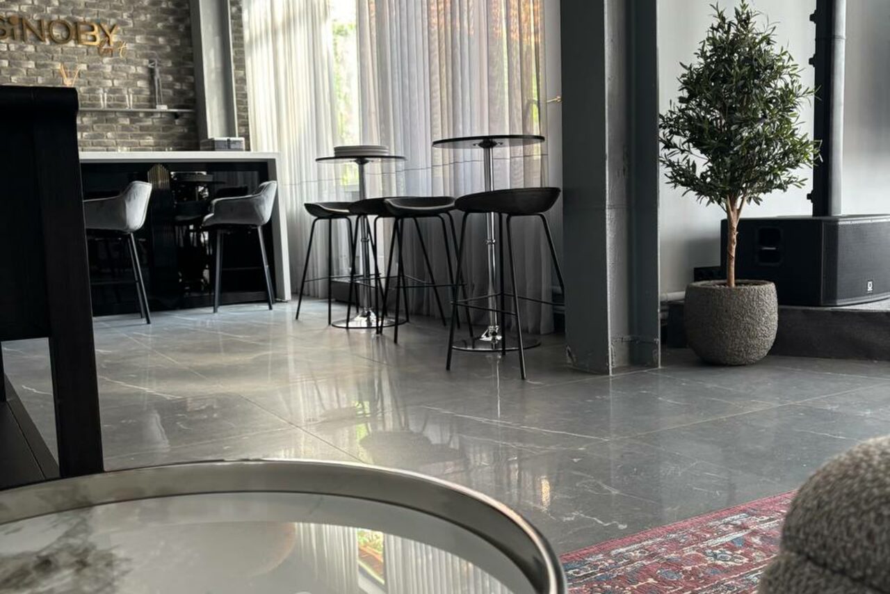 Арендовать Лофт Loftsinoby (Лофтсиноби)  для мероприятий. Барное пространство с высокими столами и стульями, столешницей из темного дерева и кирпичной стеной. Пол из серого камня, на столе стеклянный круг, рядом зеленое растение в горшке. Свет через прозрачные шторы, коврик с узором.