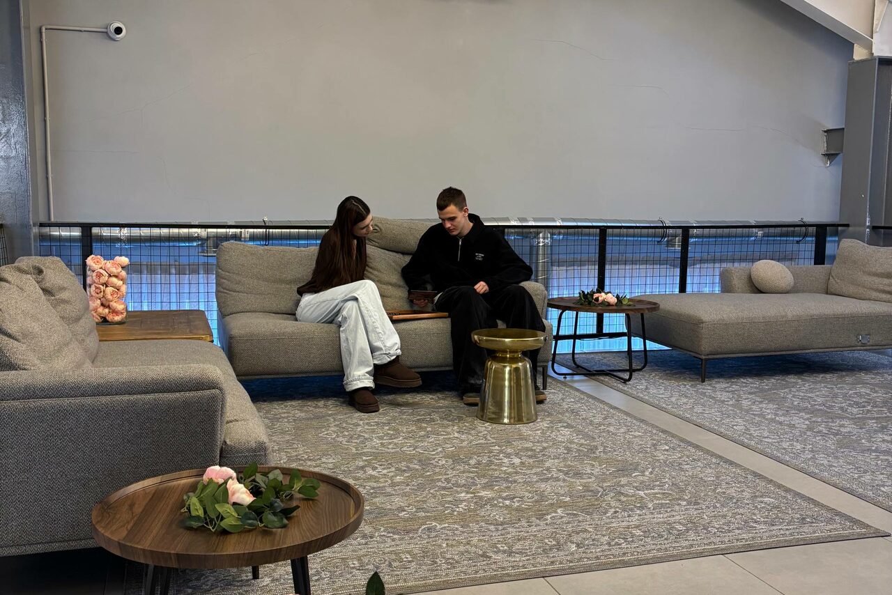 Арендовать Лофт Loftsinoby (Лофтсиноби)  для мероприятий. Современное помещение с серыми диванами и коврами. Невысокие металлические столики, на одном из которых цветочная композиция. Стены серого цвета. Металлические перила вдоль стены. Полы выложены плиткой.