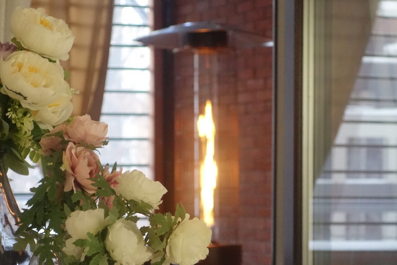 Арендовать Лофт Loftsinoby (Лофтсиноби)  для мероприятий. Цветы на переднем плане, обогреватель с открытым пламенем на фоне. Стена из красного кирпича, большие окна с жалюзи. На переднем плане белые и розовые цветы, справа видны занавески.