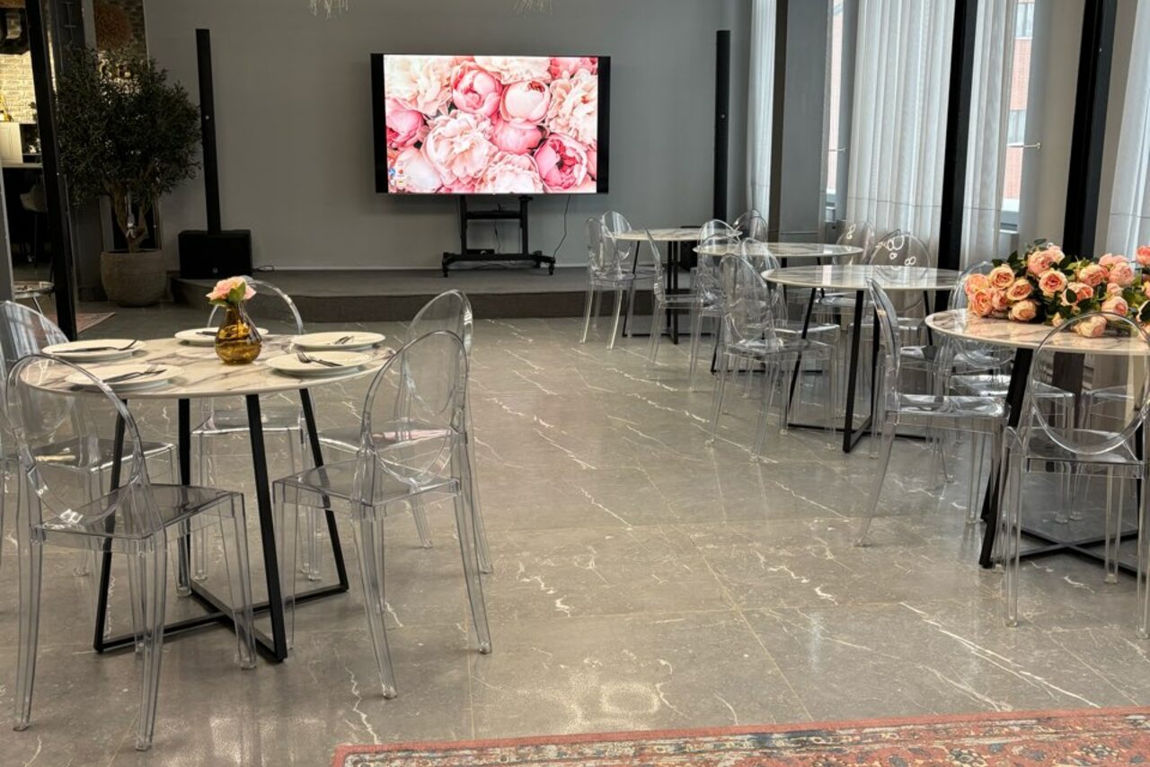 Арендовать Лофт Loftsinoby (Лофтсиноби)  для мероприятий. Пространство для мероприятий с мраморным полом, круглые столы, прозрачные стулья, ваза с цветами, большой экран с изображением цветов, окно с белыми шторами, горшок с растением.