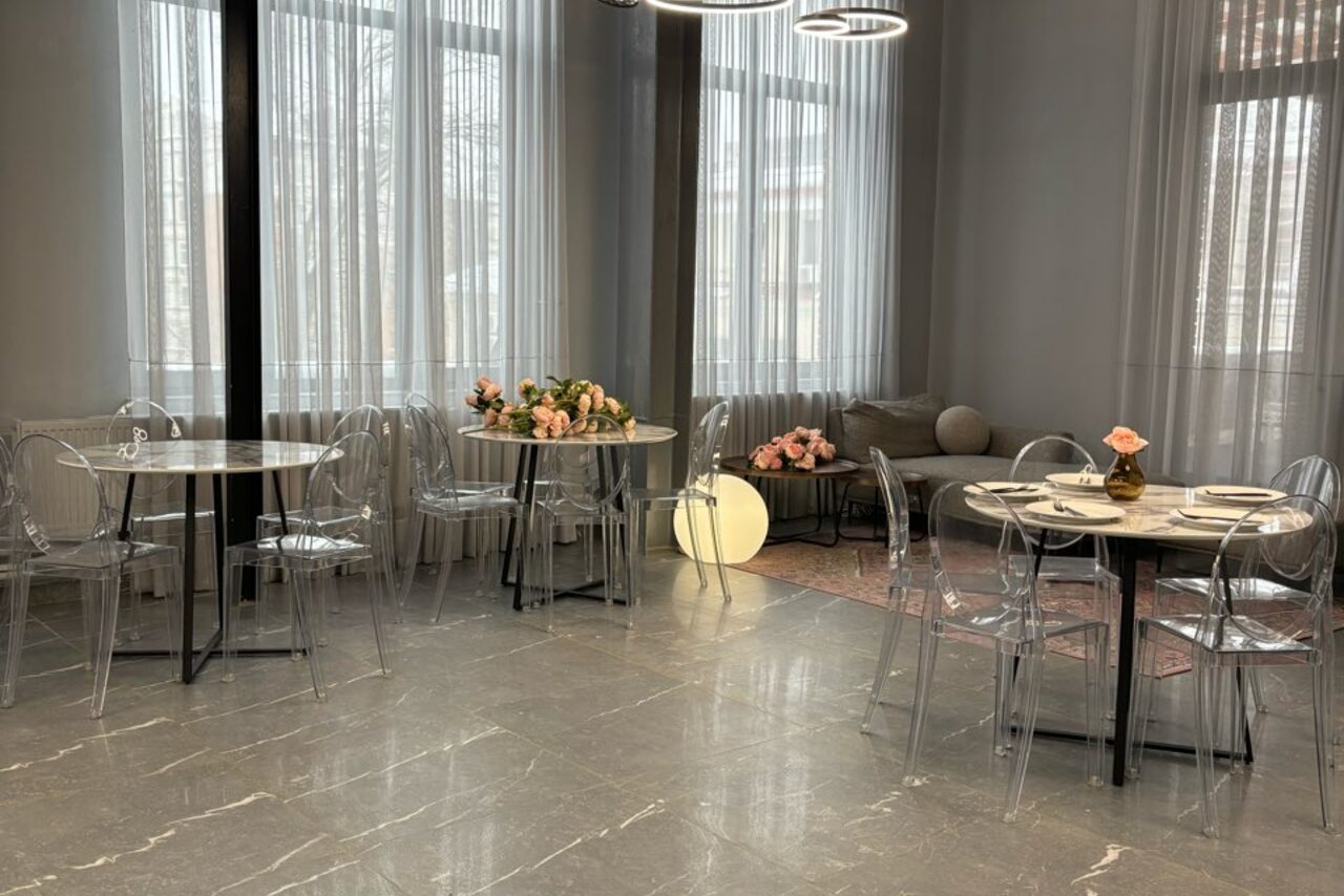 Арендовать Лофт Loftsinoby (Лофтсиноби)  для мероприятий. Современное кафе с круглыми столами и прозрачными стульями. Светлые шторы на окнах, мягкий диван в углу. Каменный пол, белые стены, центральное круглое освещение. На столах цветочные композиции.