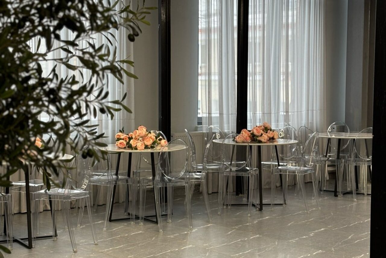 Арендовать Лофт Loftsinoby (Лофтсиноби)  для мероприятий. Современное светлое помещение с круглыми столами и прозрачными пластиковыми стульями. На столах розовые цветочные композиции. Пол мраморный, окна с белыми шторами. В левом углу растения.