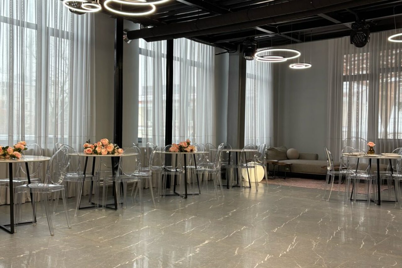 Арендовать Лофт Loftsinoby (Лофтсиноби)  для мероприятий. Современное пространство с большими окнами и занавесями, пол из серого камня с узорами. Столы круглой формы с прозрачными стульями, декорированы цветочными композициями. Круглые подвесные светильники на потолке, зона отдыха с диваном.